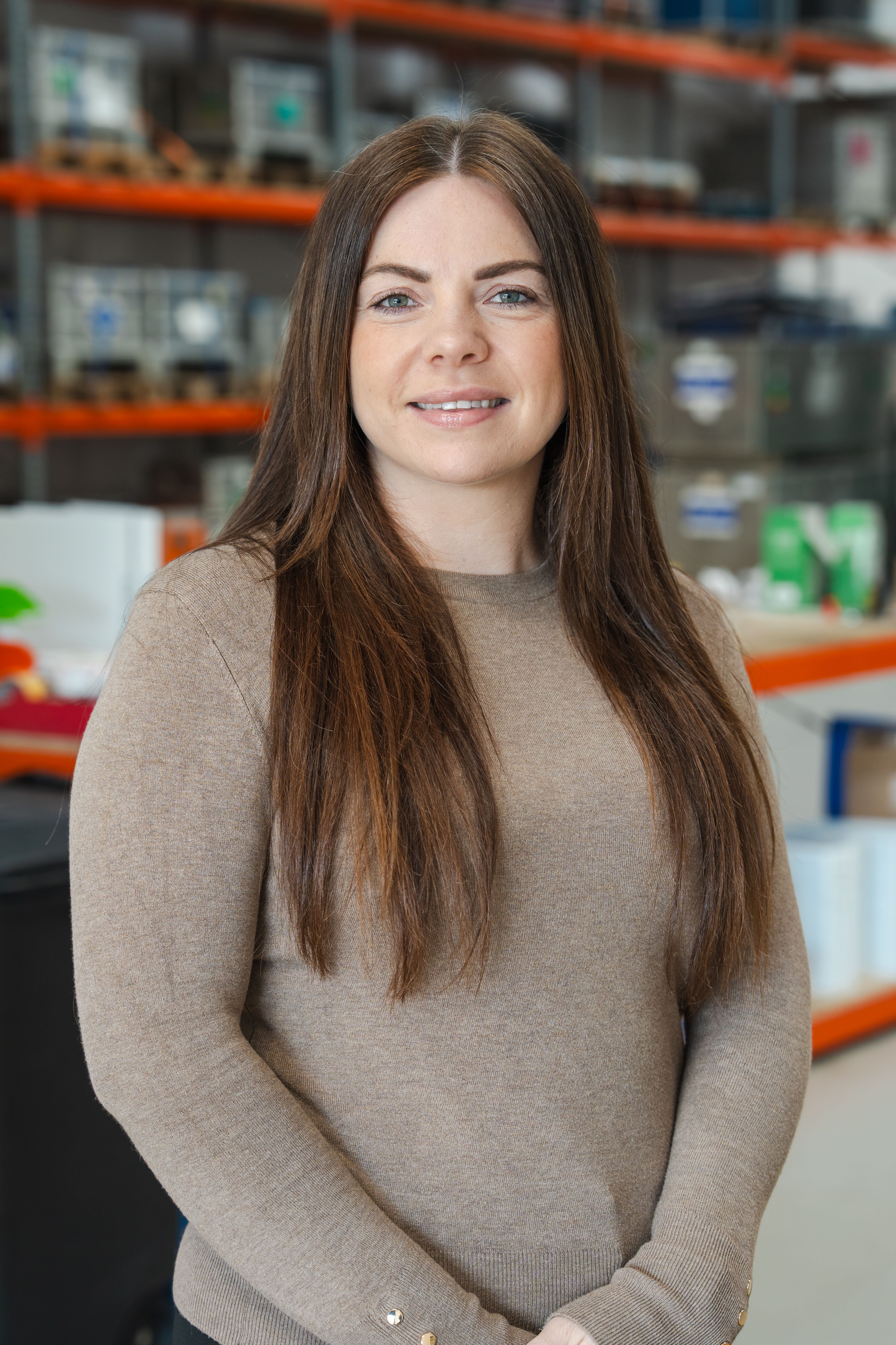 A smiling woman with long brown hair and a beige sweater stands in a blurred warehouse.