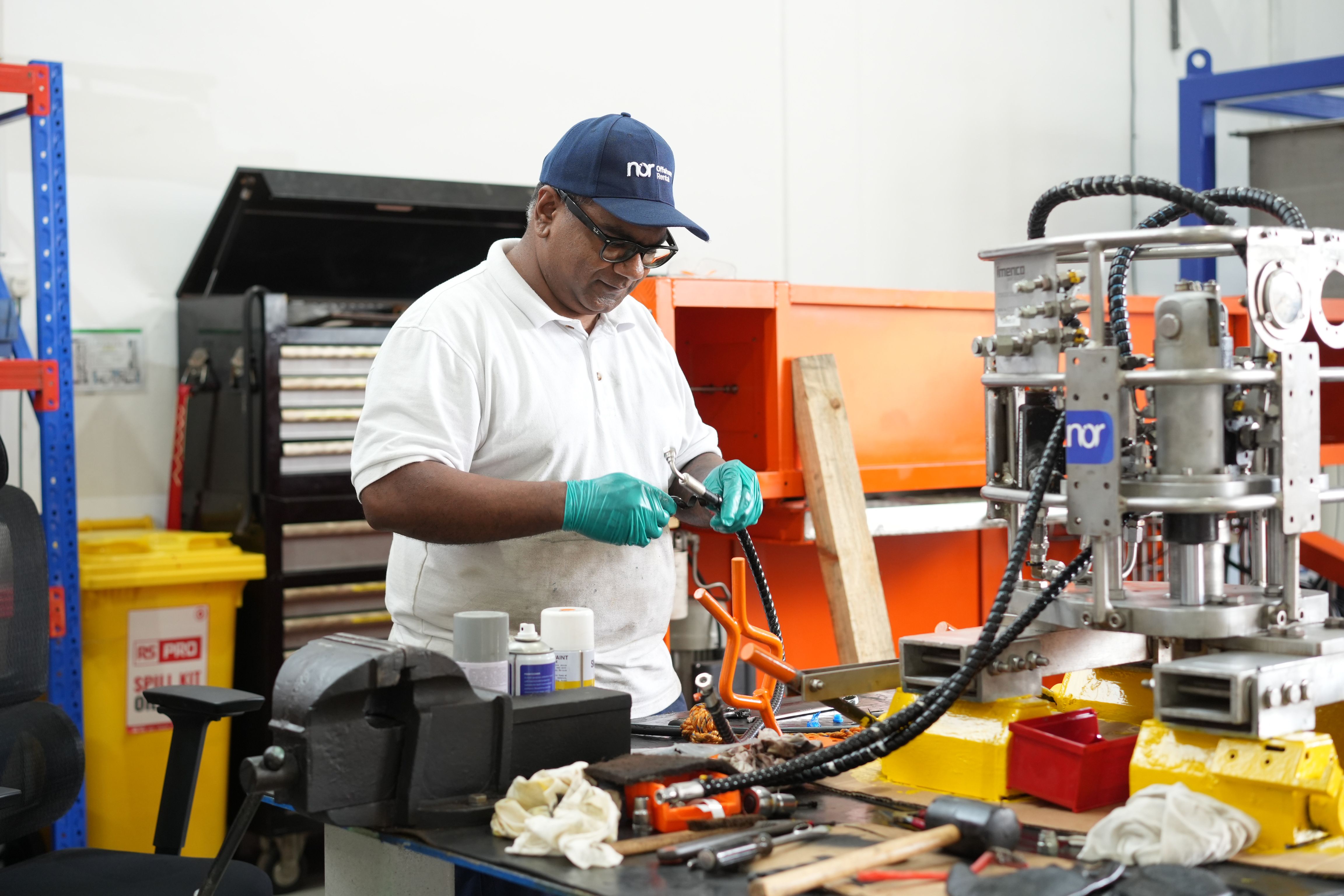 A technician wearing a Nor cap and gloves works on a mechanical part at a workbench in a workshop.