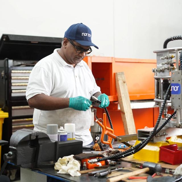 A technician wearing a Nor cap and gloves works on a mechanical part at a workbench in a workshop.