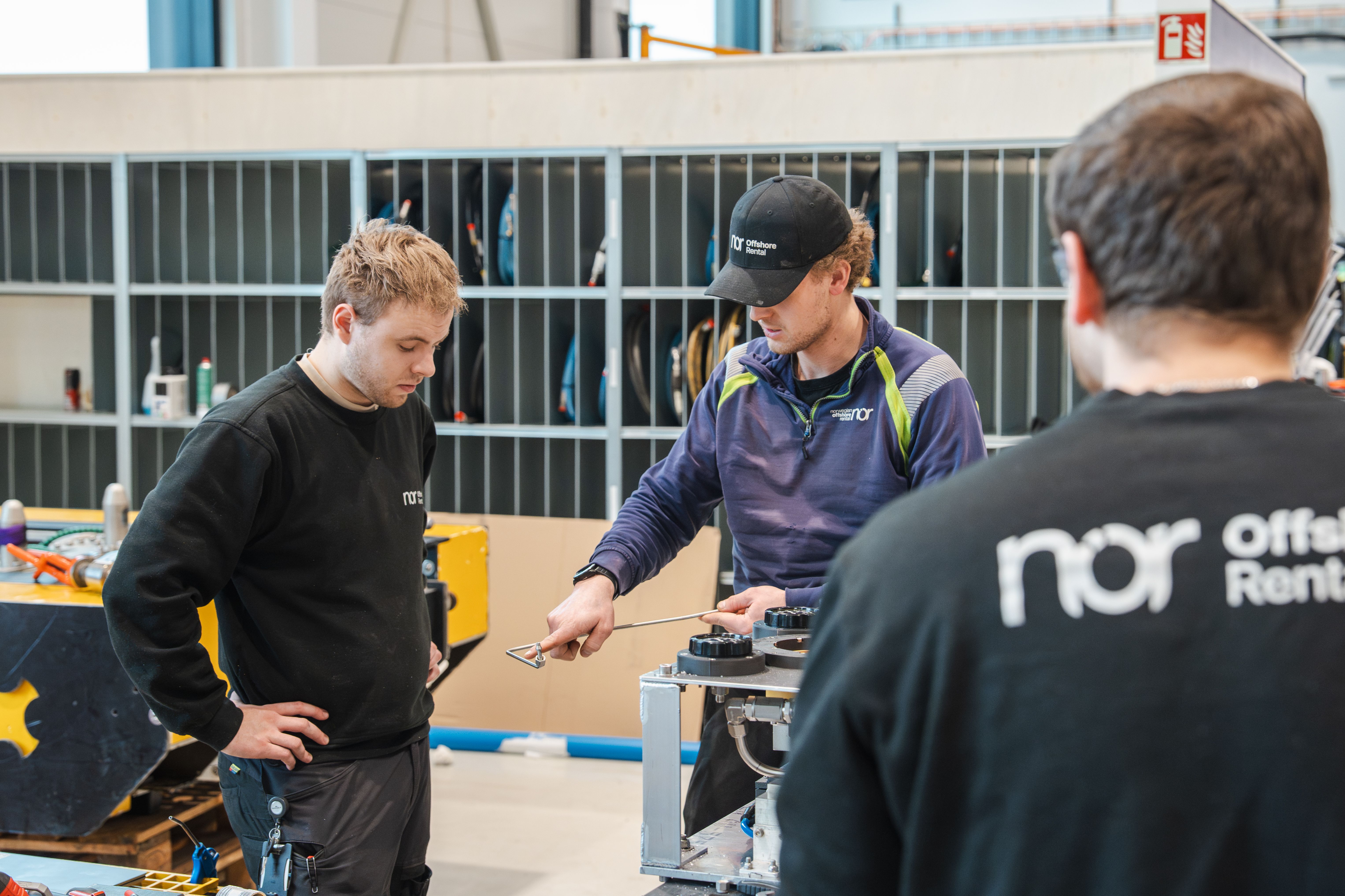 Three men in a workshop, one demonstrating on equipment while another observes.
