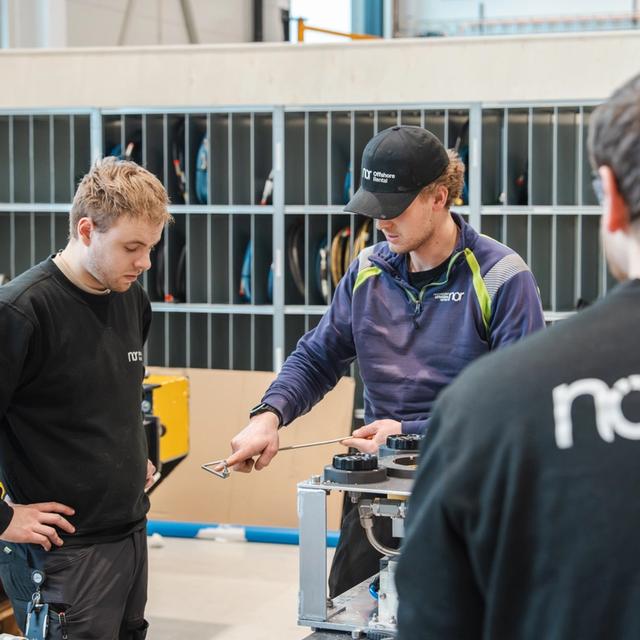 Three men in a workshop, one demonstrating on equipment while another observes.