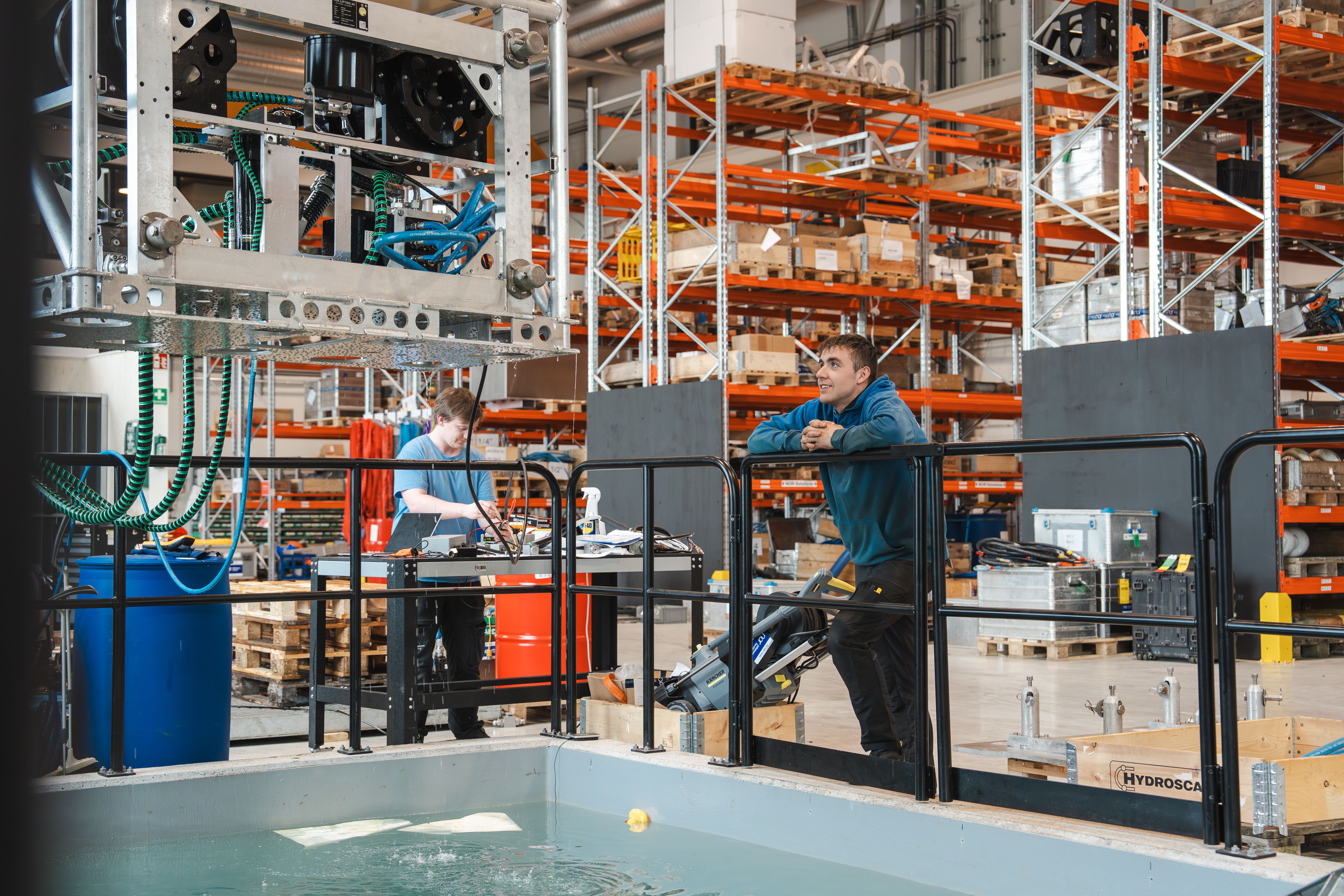 A man in a blue hoodie leans on a black railing above a water tank, looking upwards in a warehouse with orange storage shelves.