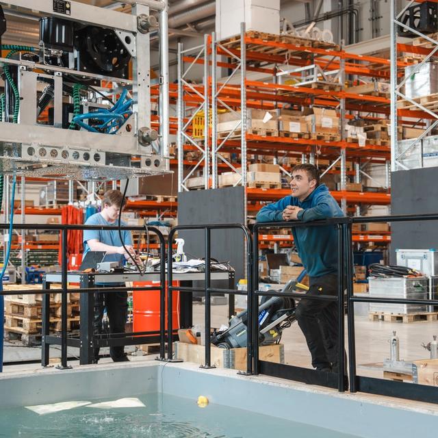 Two engineers work in a lab with a large water tank, overhead machinery, and warehouse shelves.