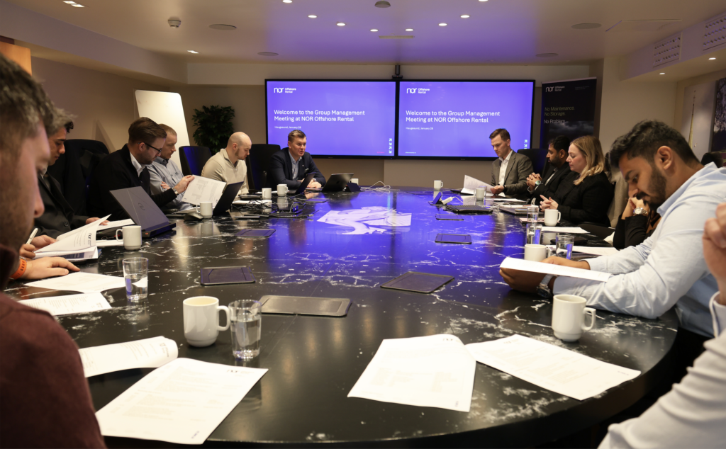 A group of people at a business meeting around a large table, with screens in the background displaying "Group Management Meeting at NOR Offshore Rental."