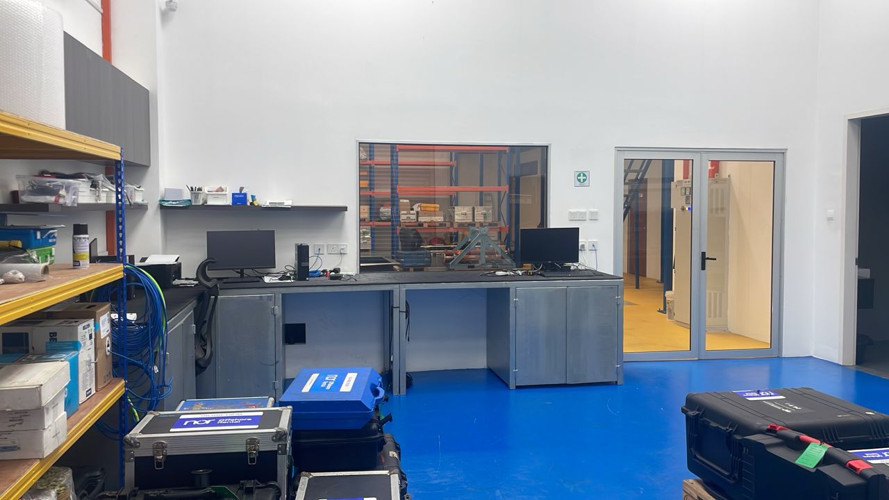 An organized lab or workshop with a blue floor, workbench, shelves, and a window to a storage area.