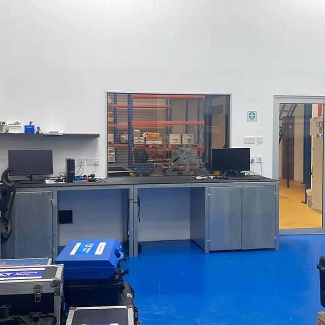 An organized lab or workshop with a blue floor, workbench, shelves, and a window to a storage area.