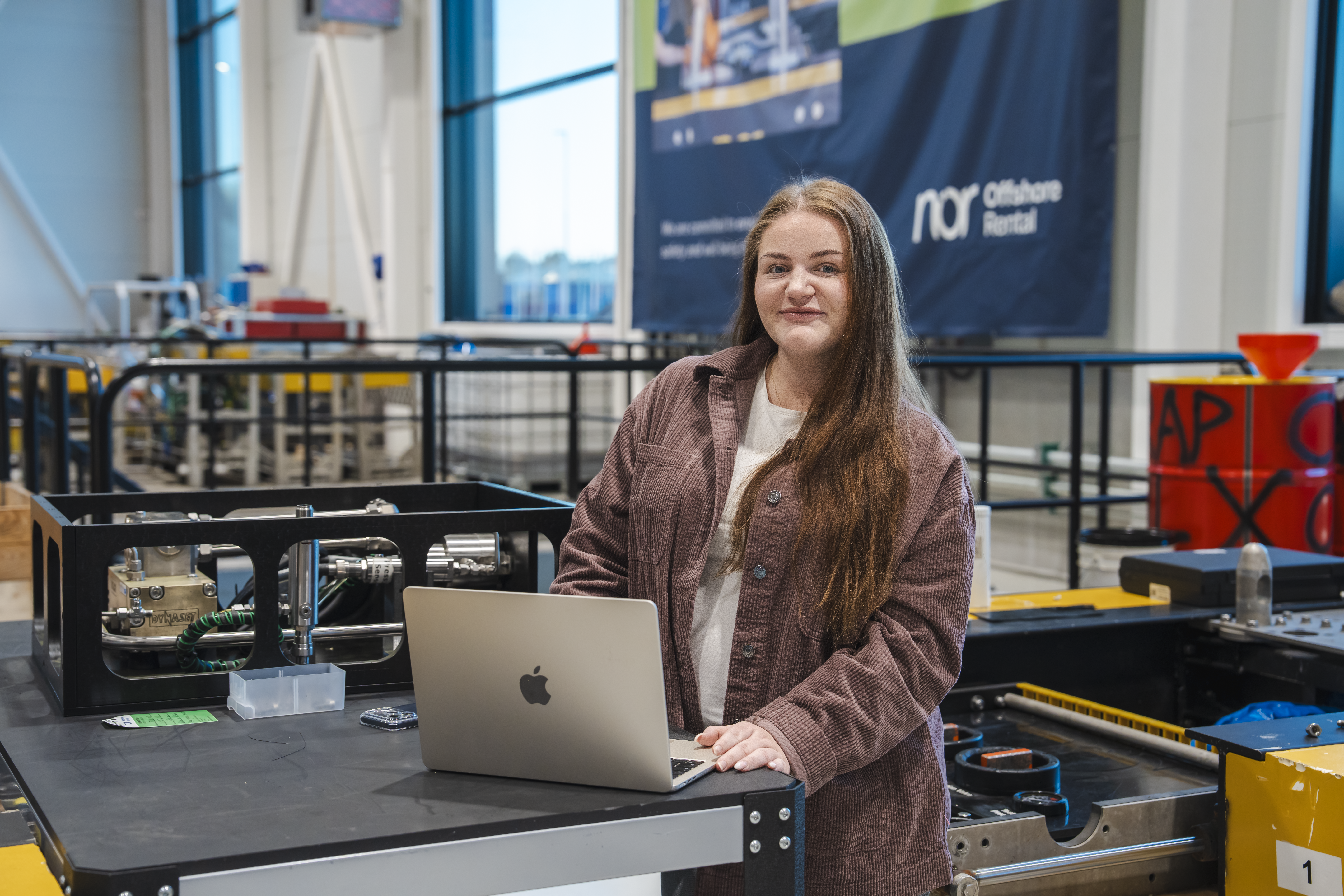A smiling young woman stands at a workbench with a laptop and industrial machinery in a workshop.