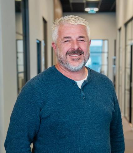 A smiling man with a gray beard wearing a blue long-sleeved shirt.