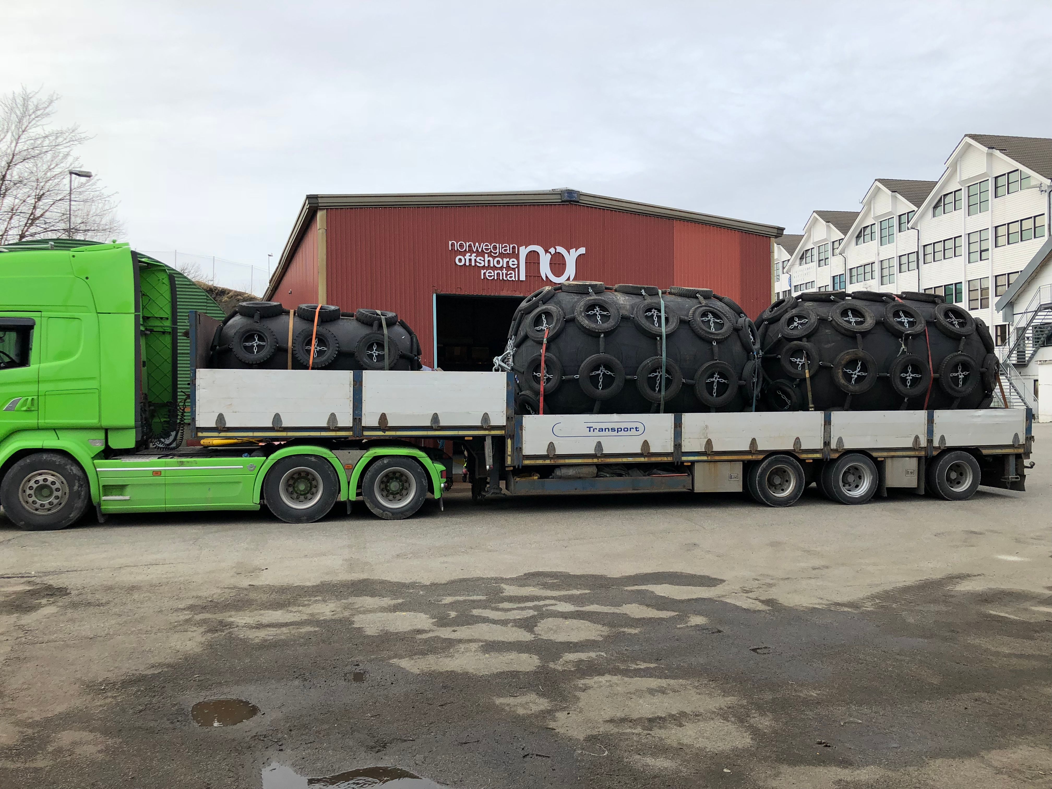 A green semi-truck carries five large, tire-covered marine fenders on a flatbed trailer in front of a "Norwegian Offshore Rental" building.