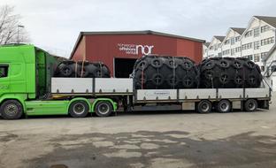 A green semi-truck carries five large, tire-covered marine fenders on a flatbed trailer in front of a "Norwegian Offshore Rental" building.