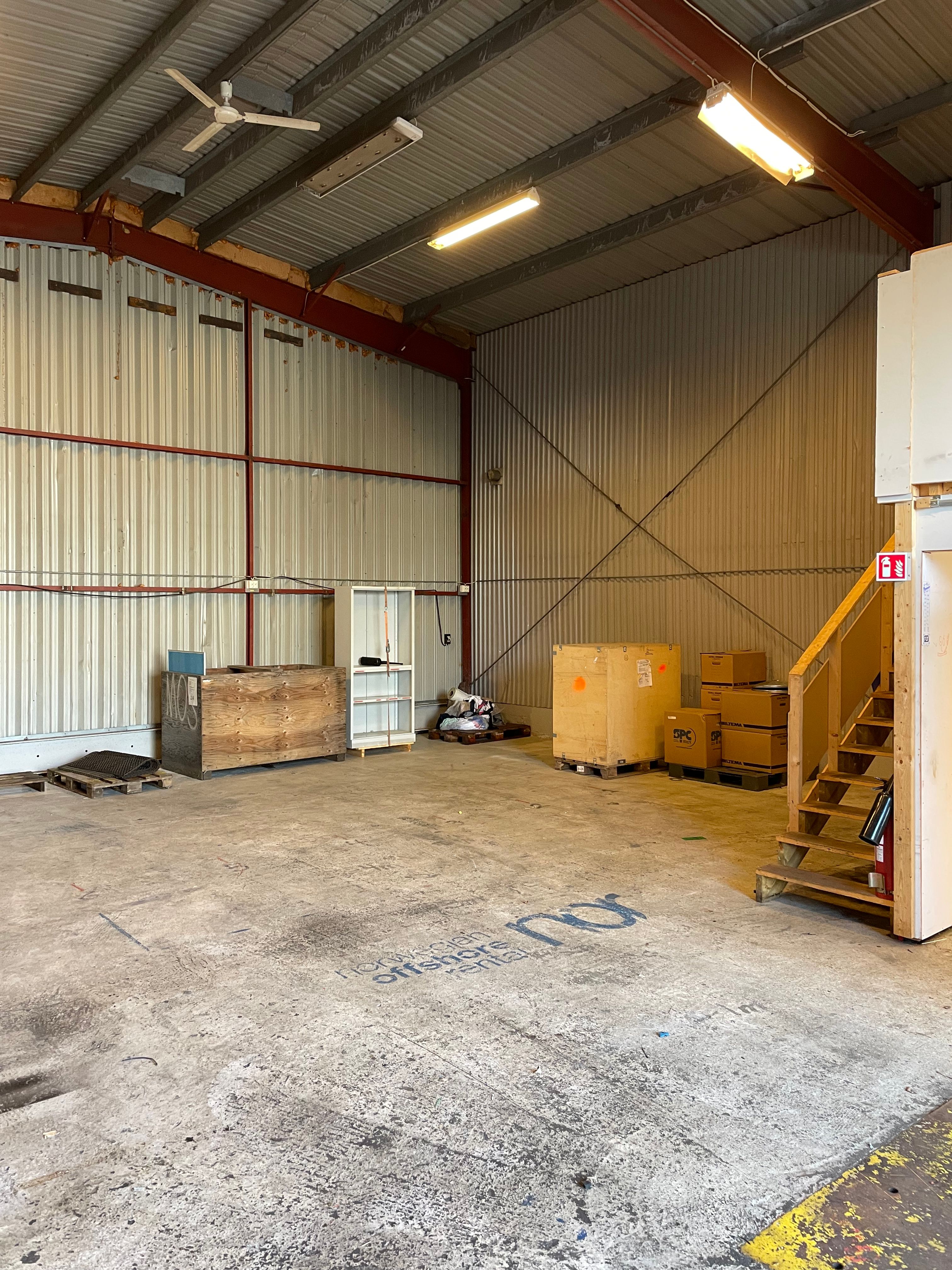 A spacious warehouse interior with a high metal ceiling, concrete floor, wooden crates, stairs, and a "Norwegian Offshore Rental" logo.