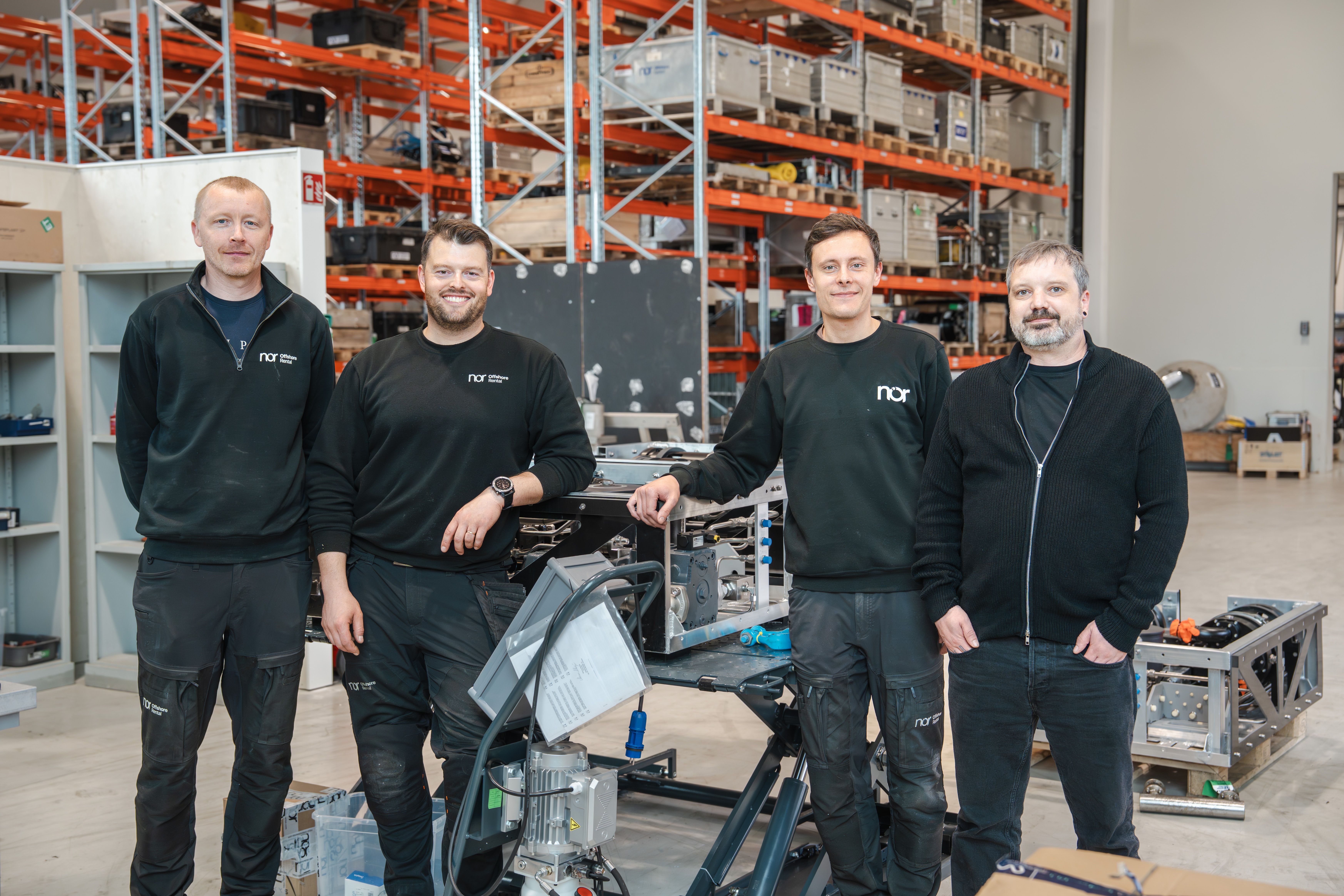 Four NOR team members standing in a workshop and warehouse environment with technical equipment and offshore project assemblies in the background.