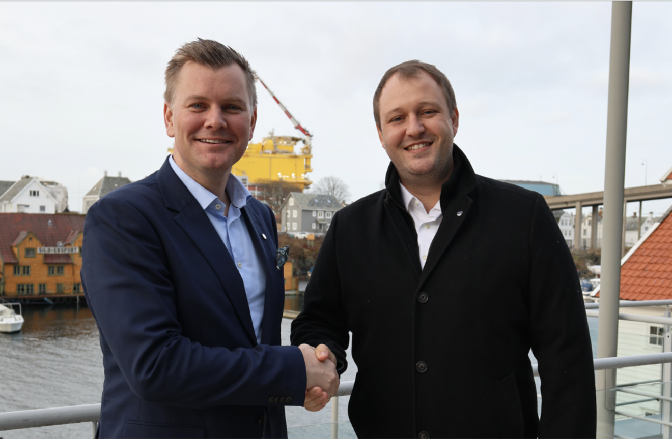 Two smiling men shake hands on a waterfront with buildings and a yellow industrial structure in the background.