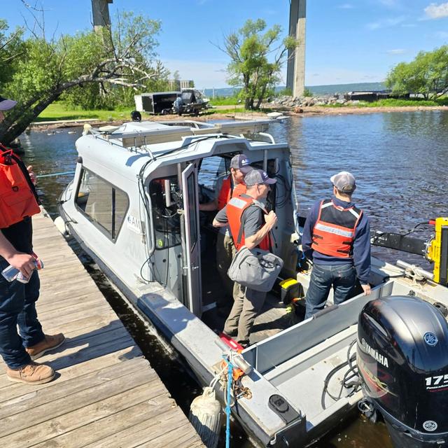 A team in life vests loads surveying equipment onto a boat docked on a sunny day.