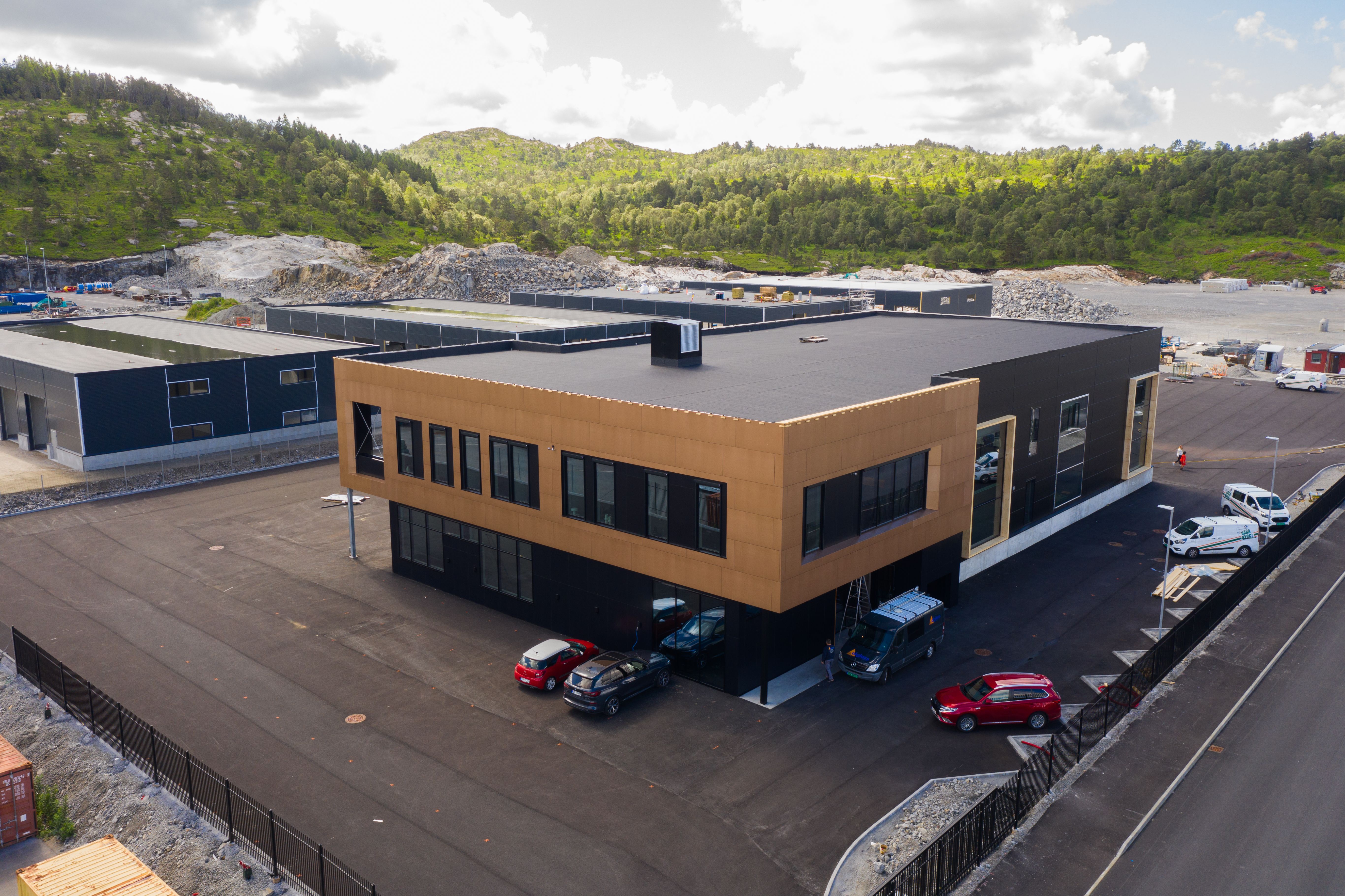 A modern two-story building with black and light brown paneling, a parking lot with cars, and industrial structures against forested hills and a construction site.