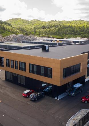 A modern two-story building with black and light brown paneling, a parking lot with cars, and industrial structures against forested hills and a construction site.