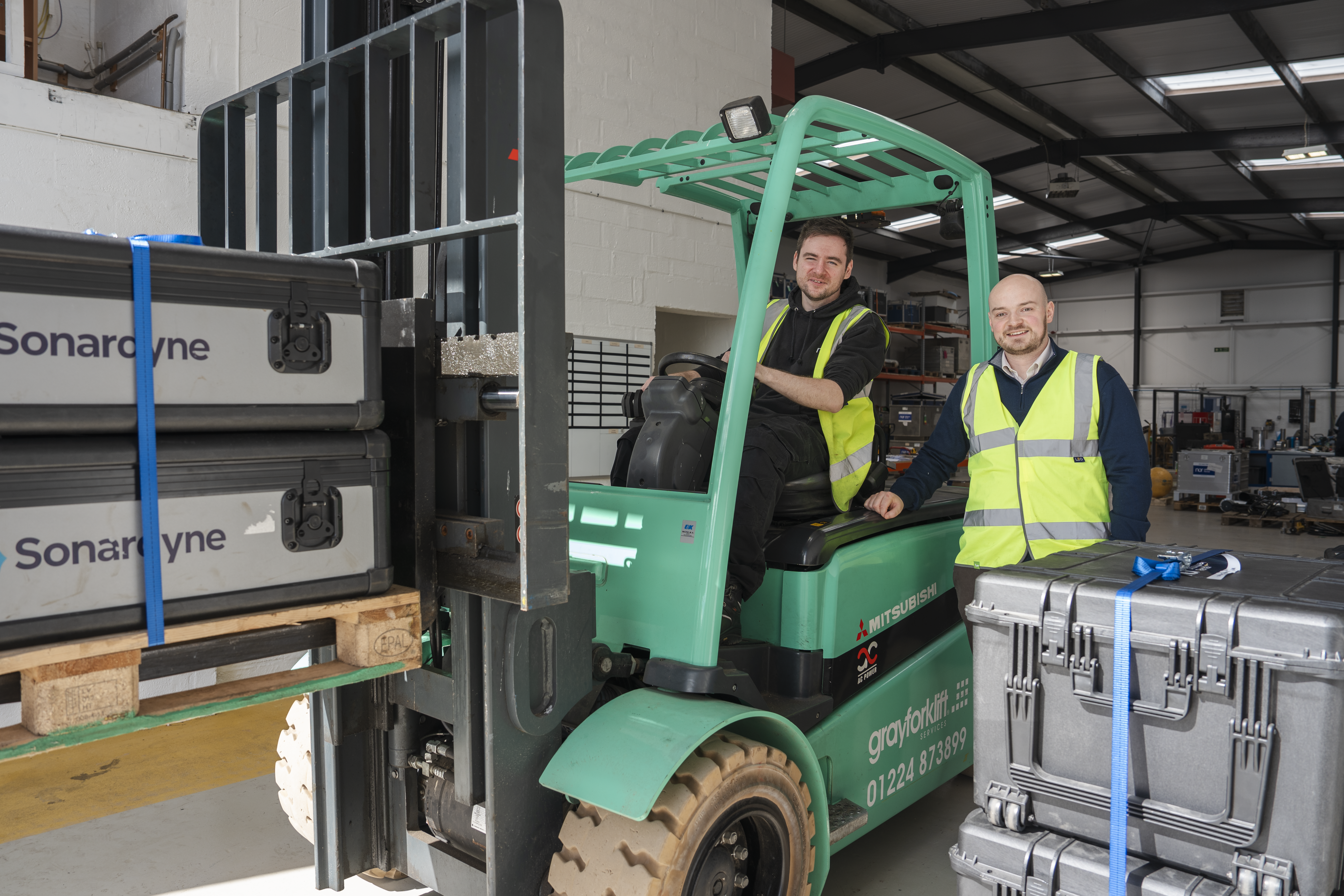 Two men in high-vis vests with a green forklift moving Sonardyne branded cases in a warehouse.