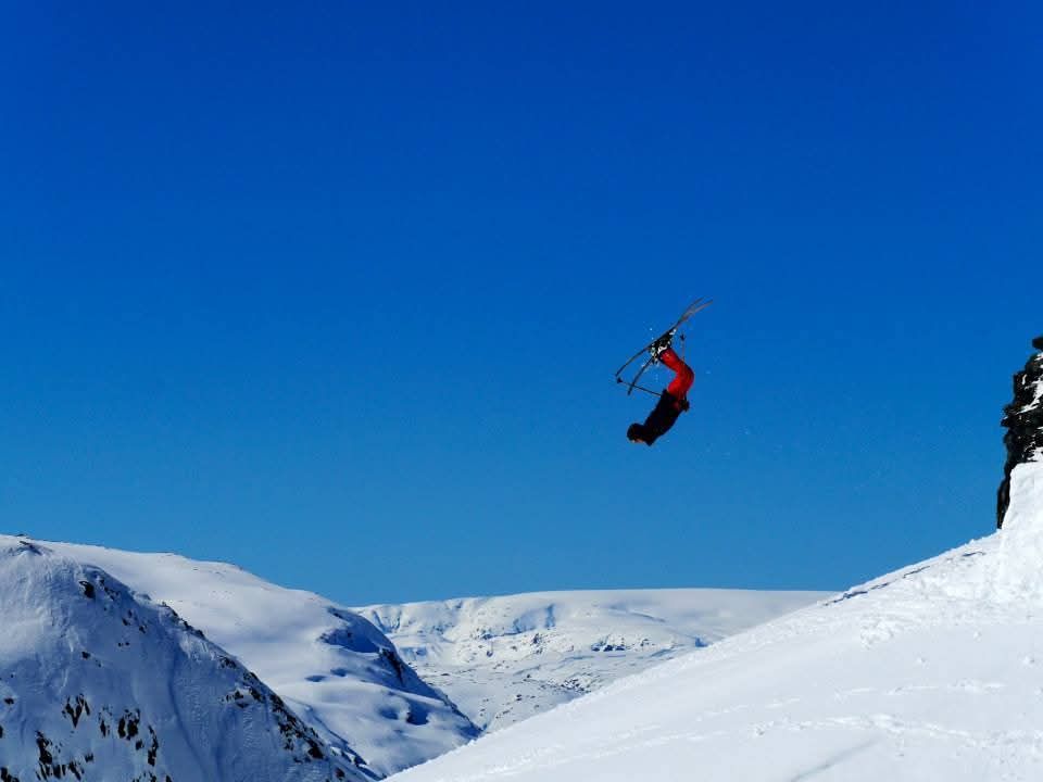 A skier performs a backflip in mid-air over snow-covered mountains under a clear blue sky.