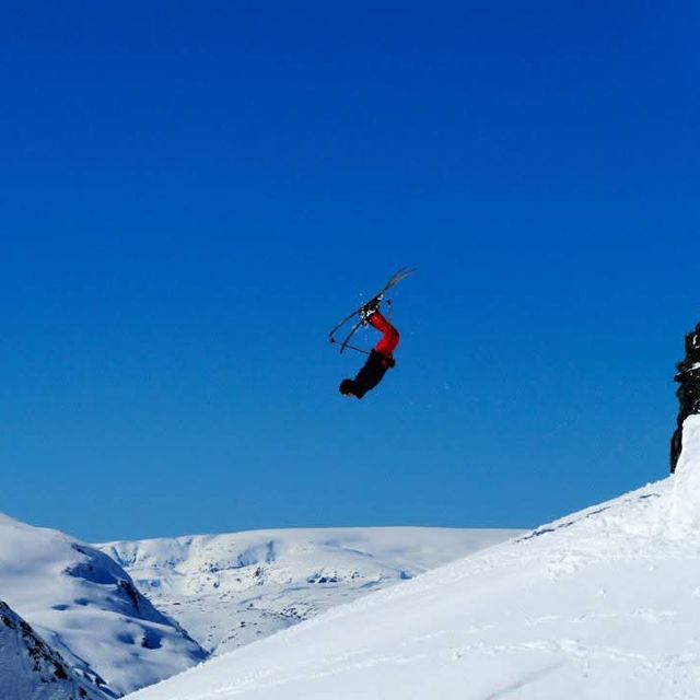 A skier performs a backflip in mid-air over snow-covered mountains under a clear blue sky.