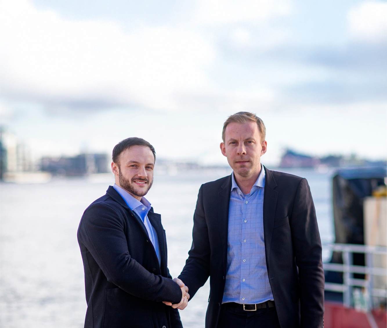 Two men in business attire shaking hands outdoors with a body of water and buildings in the background.