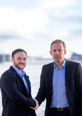 Two men in business attire shaking hands outdoors with a body of water and buildings in the background.