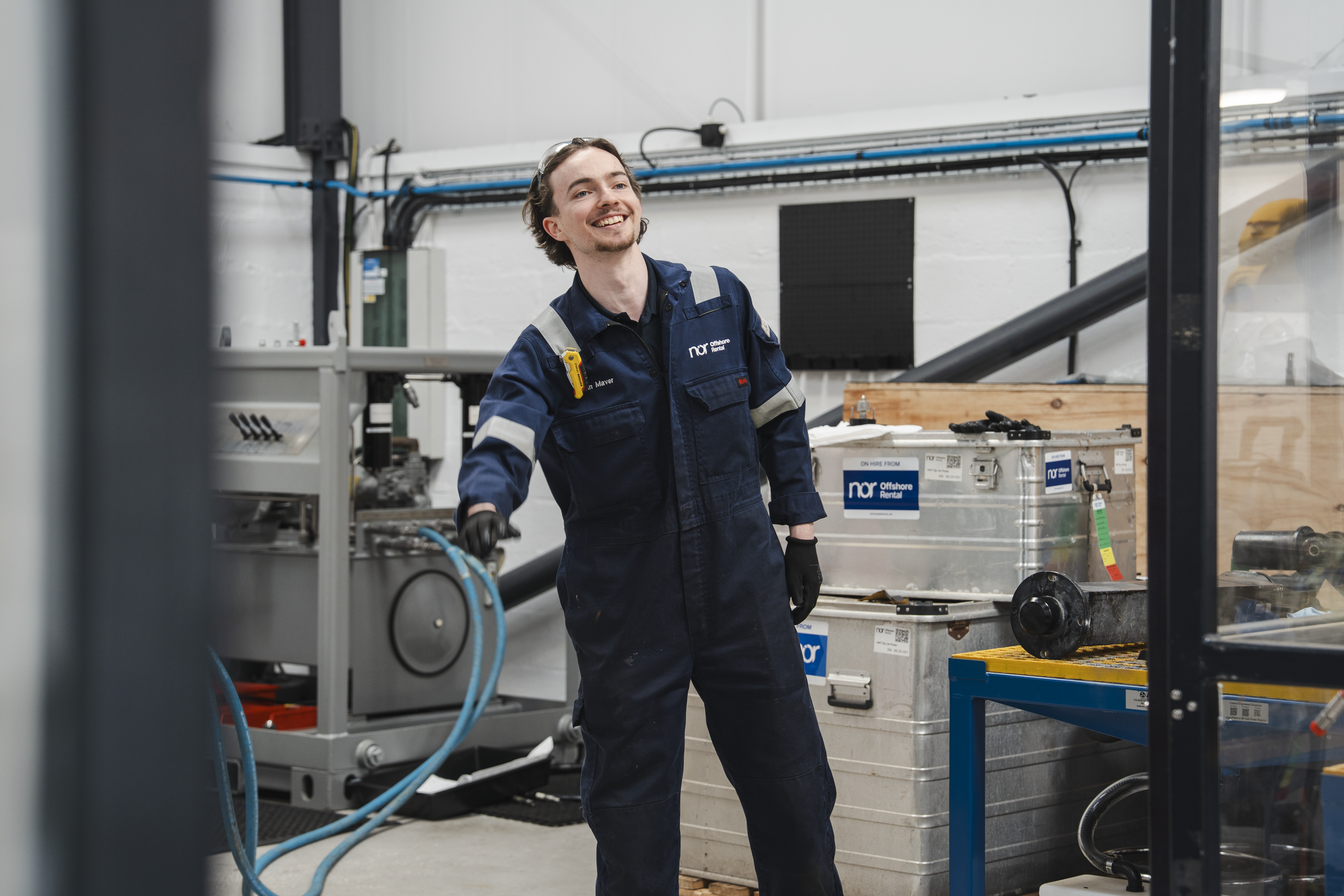 A smiling man in a blue NOR jumpsuit holding a hose in a workshop.