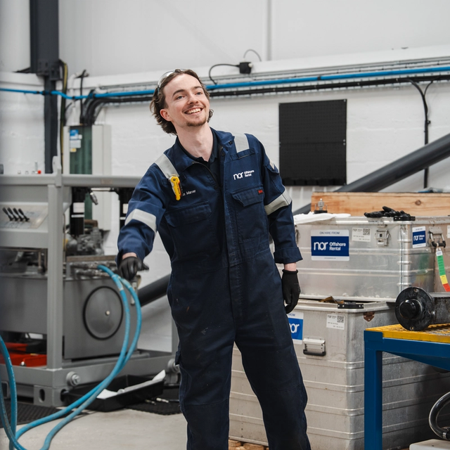 A smiling man in a blue NOR jumpsuit holding a hose in a workshop.