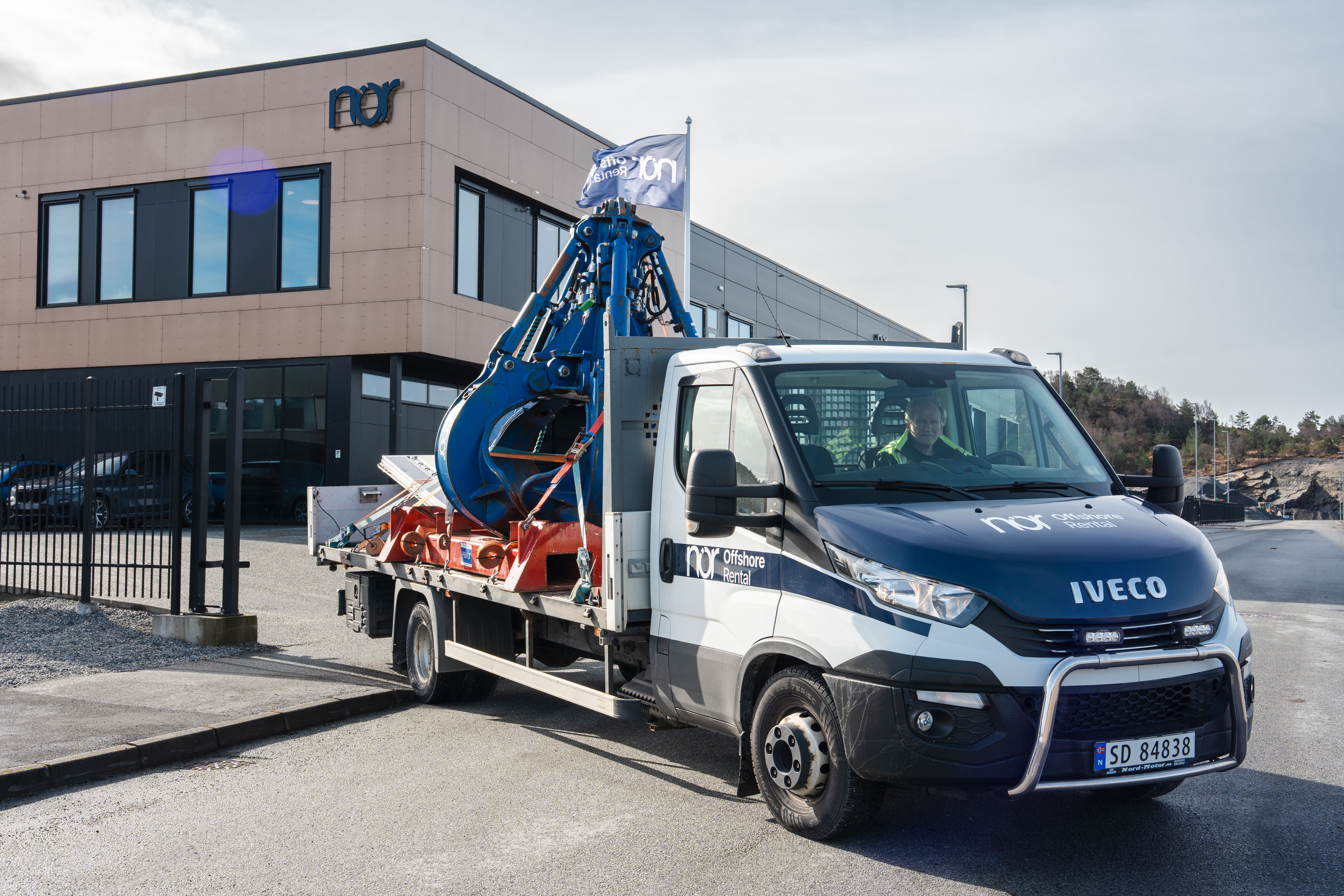 NOR Offshore branded Iveco truck carrying large industrial machinery in front of a building.