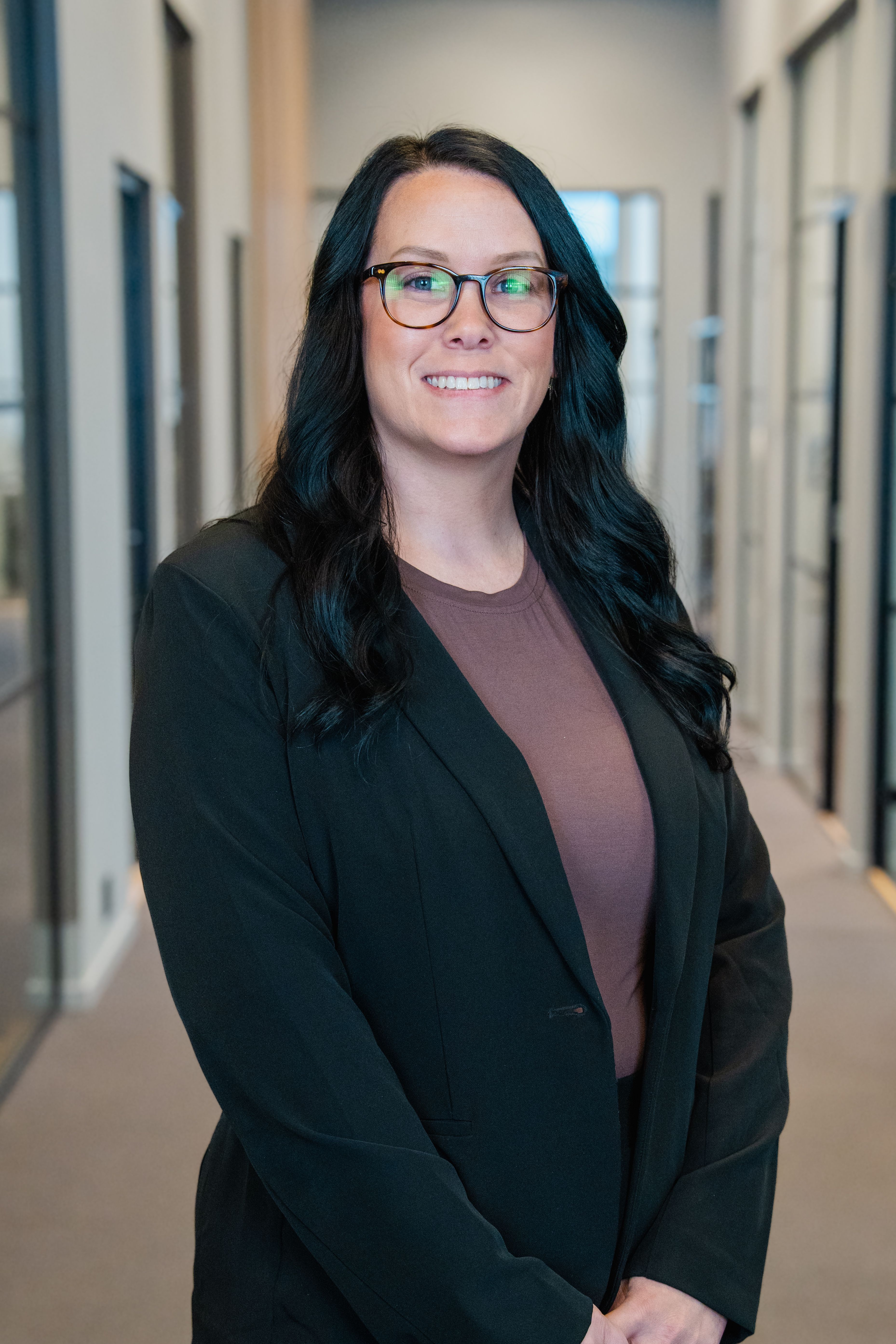 A smiling woman with long dark hair and glasses wears a dark blazer in a modern office hallway.