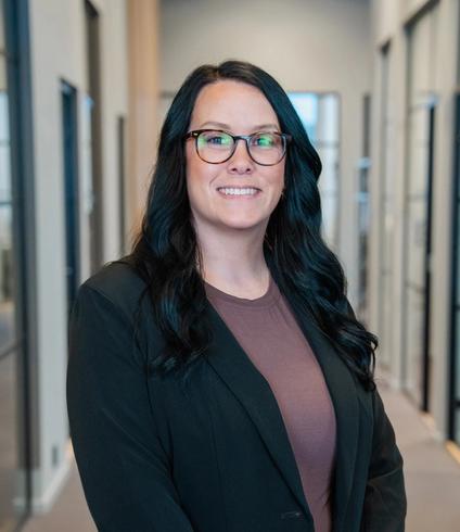 A smiling woman with long dark hair and glasses wears a dark blazer in a modern office hallway.