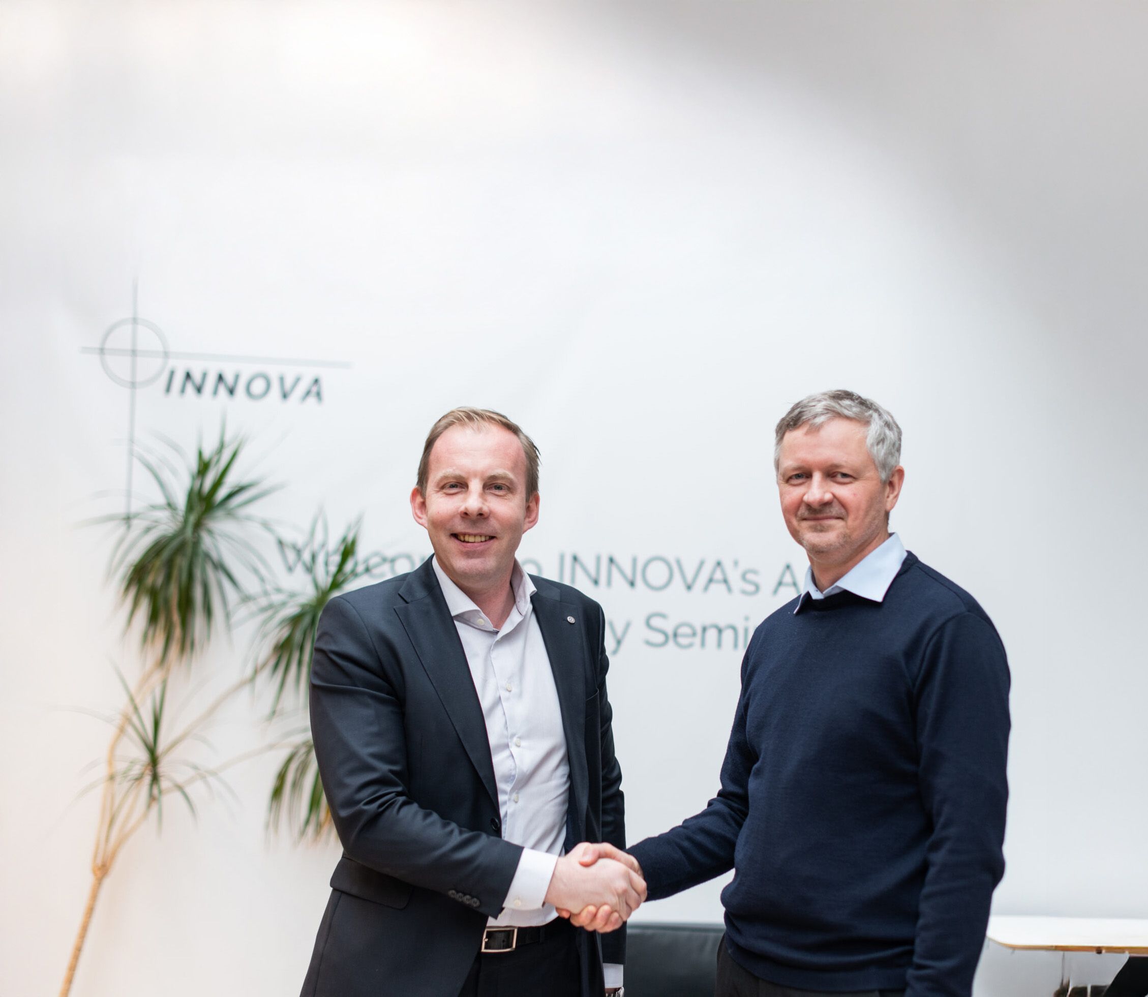 Two smiling men shake hands in an office with an Innova logo on the wall.