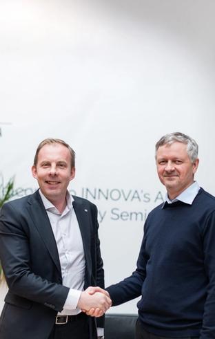Two smiling men shake hands in an office with an Innova logo on the wall.