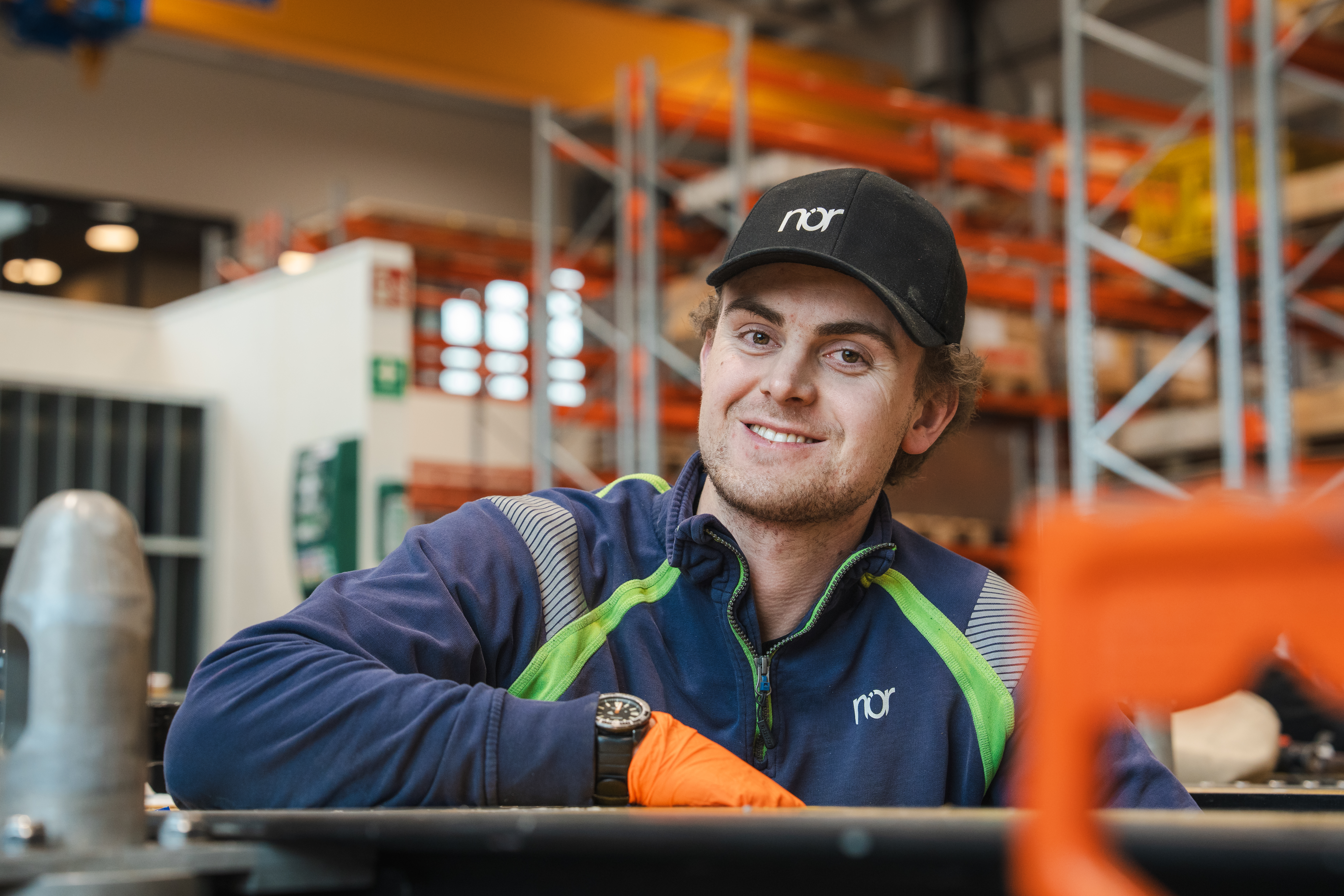 A smiling young man wearing a "nor" branded cap and uniform with an orange glove in a warehouse.