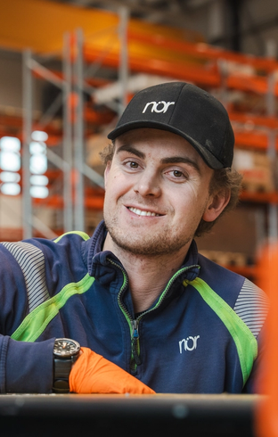 A smiling young man wearing a "nor" branded cap and uniform with an orange glove in a warehouse.