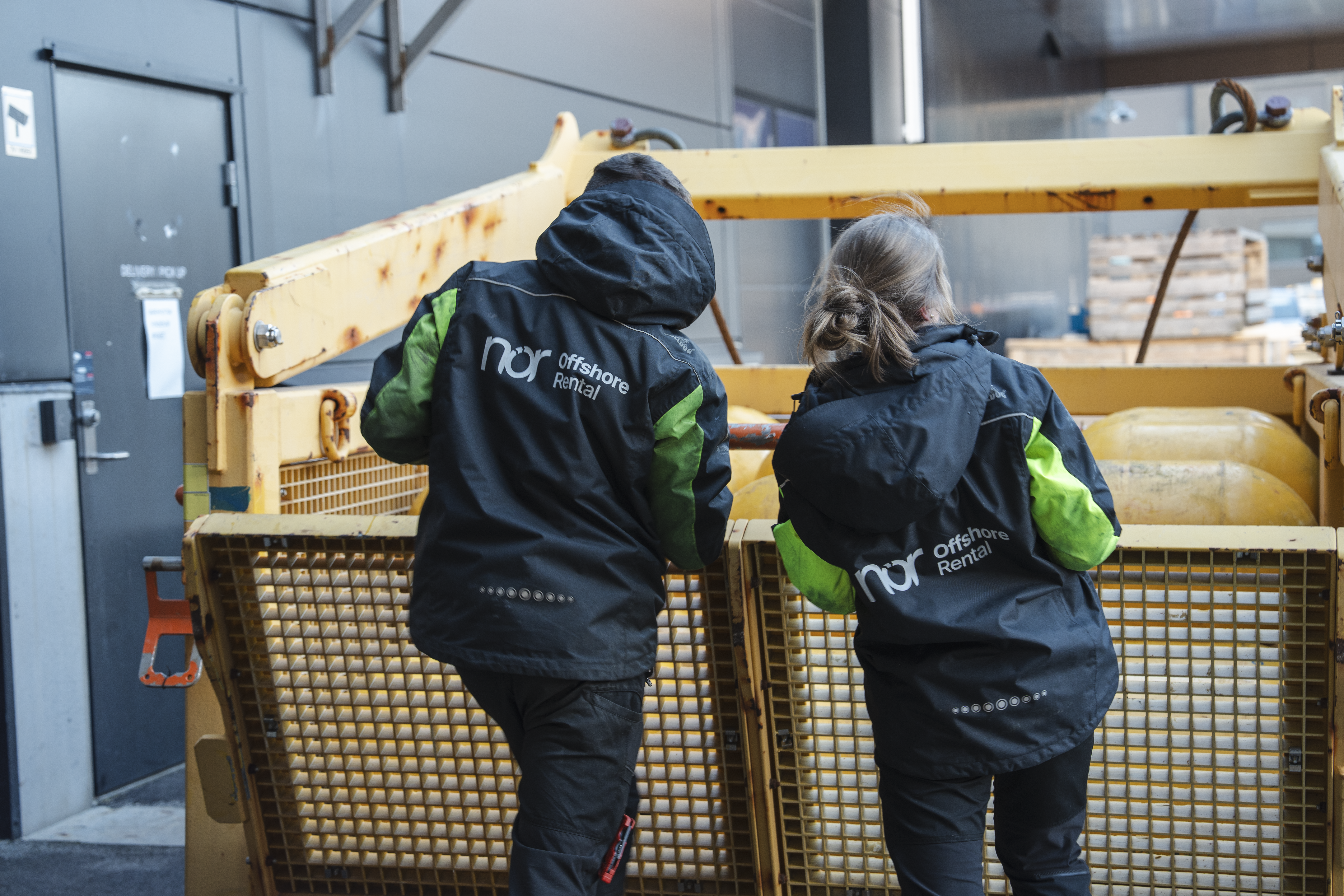 Two people in nor Offshore Rental jackets, seen from behind, stand by a large yellow industrial container with buoyant objects inside.