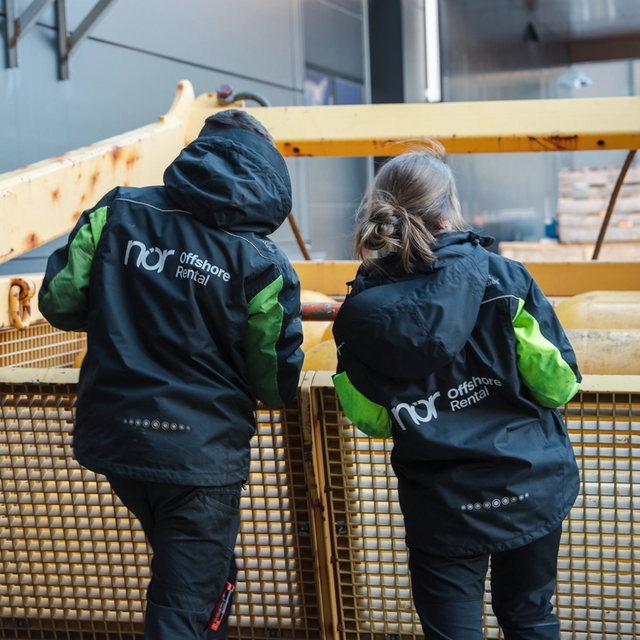 Two people in nor Offshore Rental jackets, seen from behind, stand by a large yellow industrial container with buoyant objects inside.