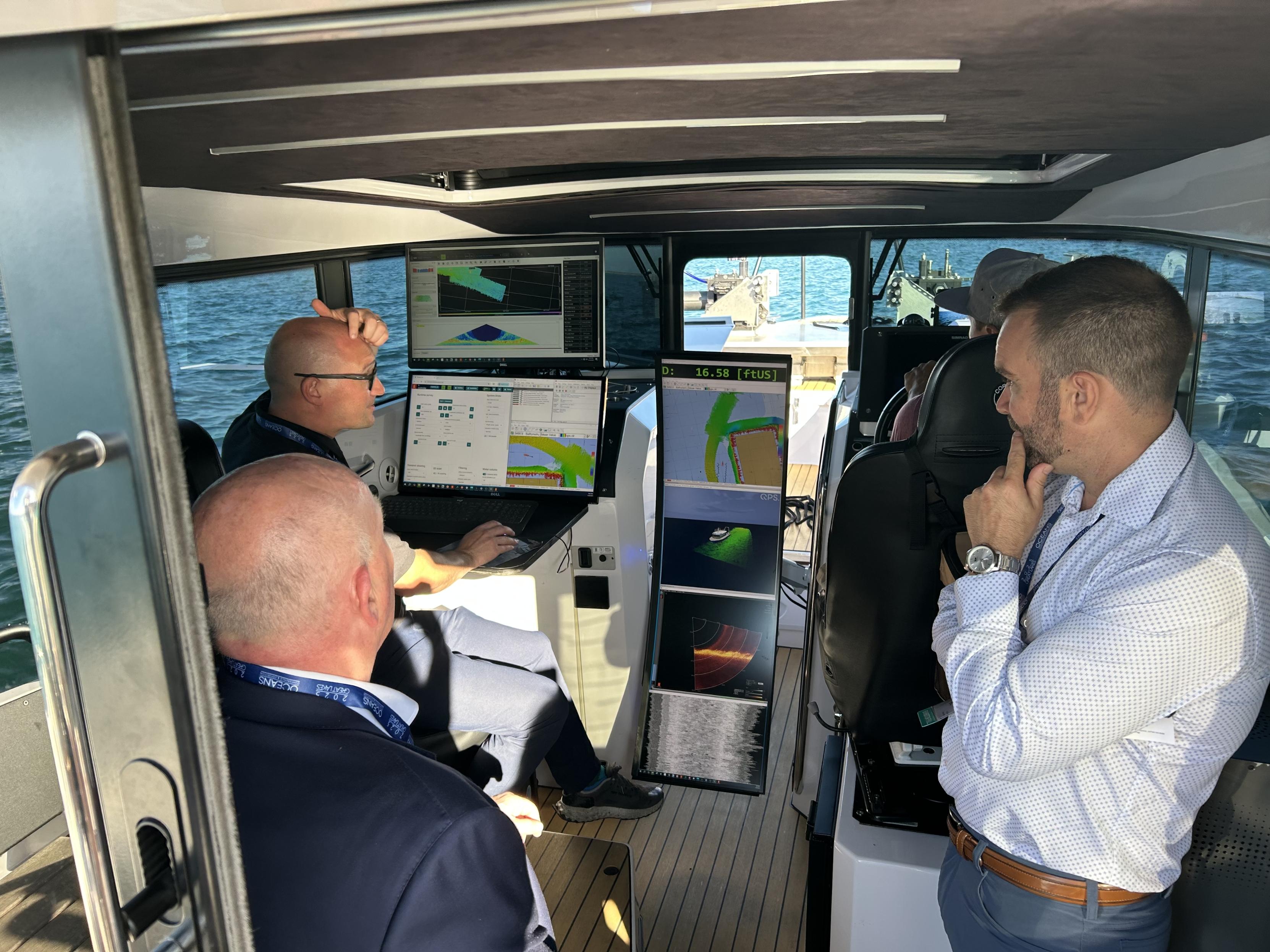 Three men in a boat cabin monitoring multiple screens displaying navigational data.
