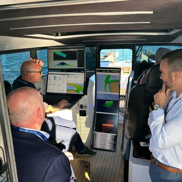 Three men in a boat cabin monitoring multiple screens displaying navigational data.