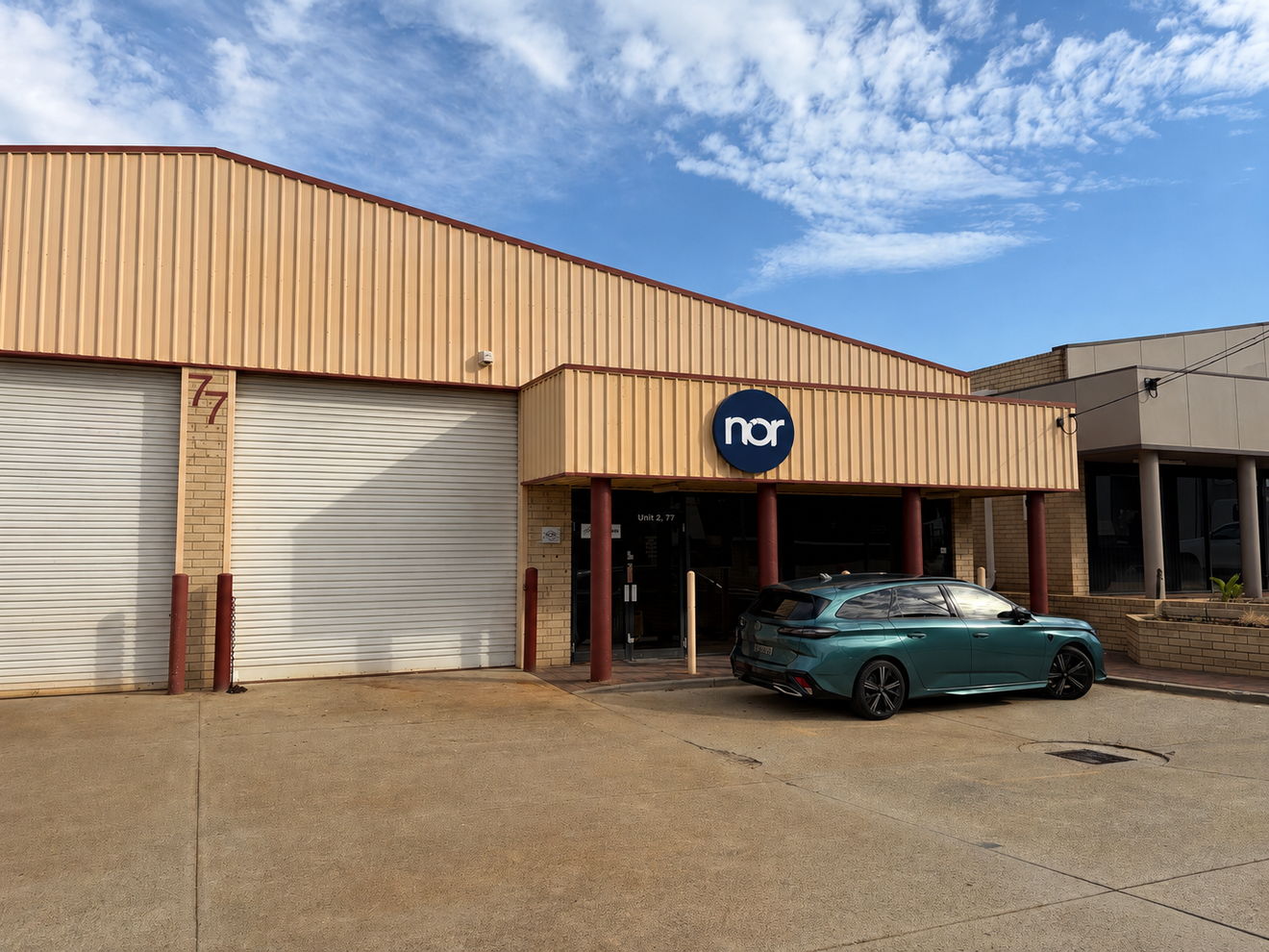 A light tan commercial building with two roll-up garage doors, a "nor" sign over the main entrance, and a dark green car parked out front.