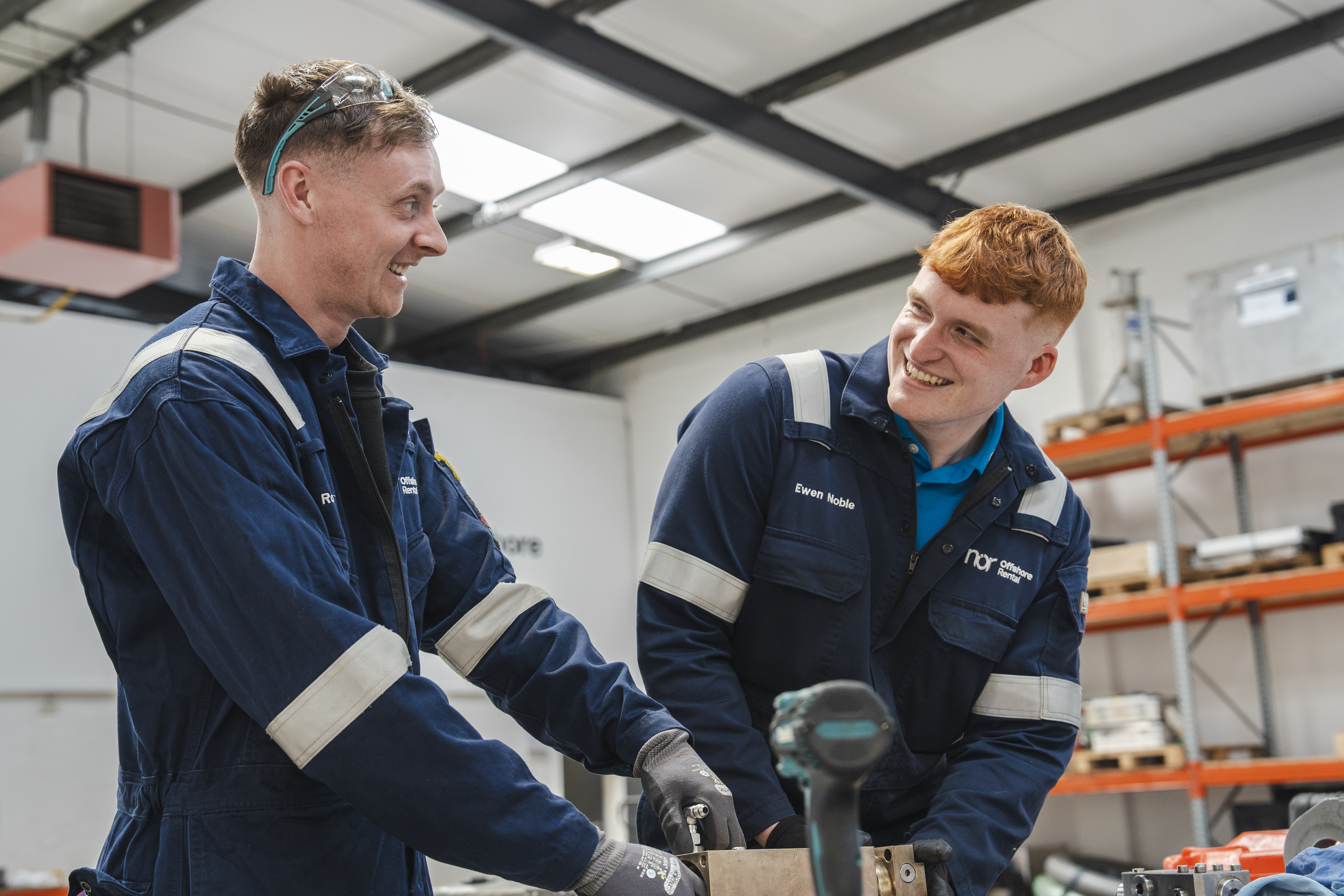 Two smiling men in blue overalls and safety gear working in a workshop.