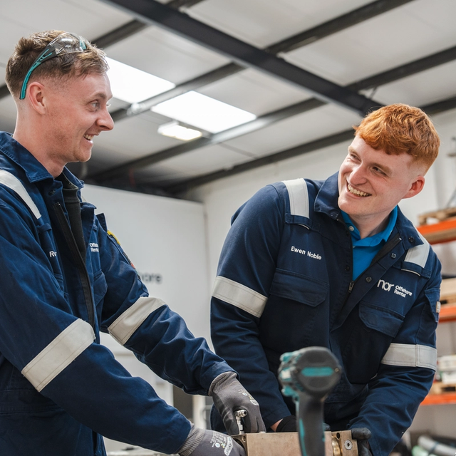 Two smiling men in blue overalls and safety gear working in a workshop.