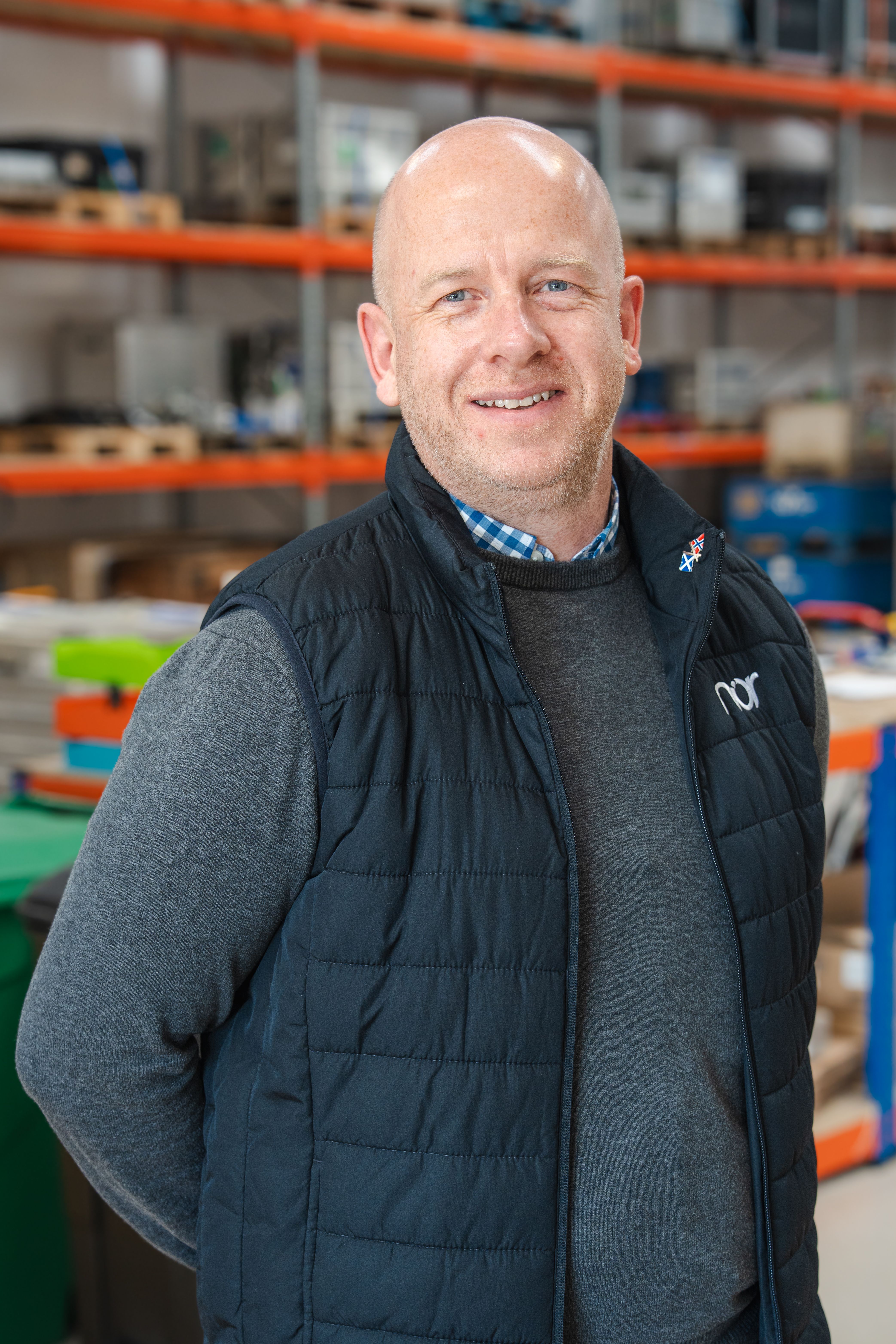 A smiling bald man in a dark vest and sweater stands in a warehouse.