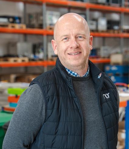 A smiling bald man in a dark vest and sweater stands in a warehouse.