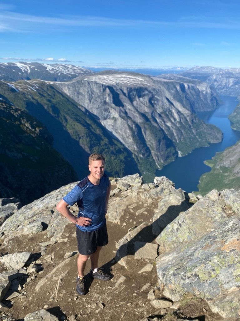 A man stands on a rocky mountain peak overlooking a scenic fjord and steep mountains.