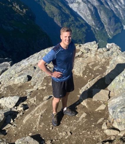 A man stands on a rocky mountain peak overlooking a scenic fjord and steep mountains.