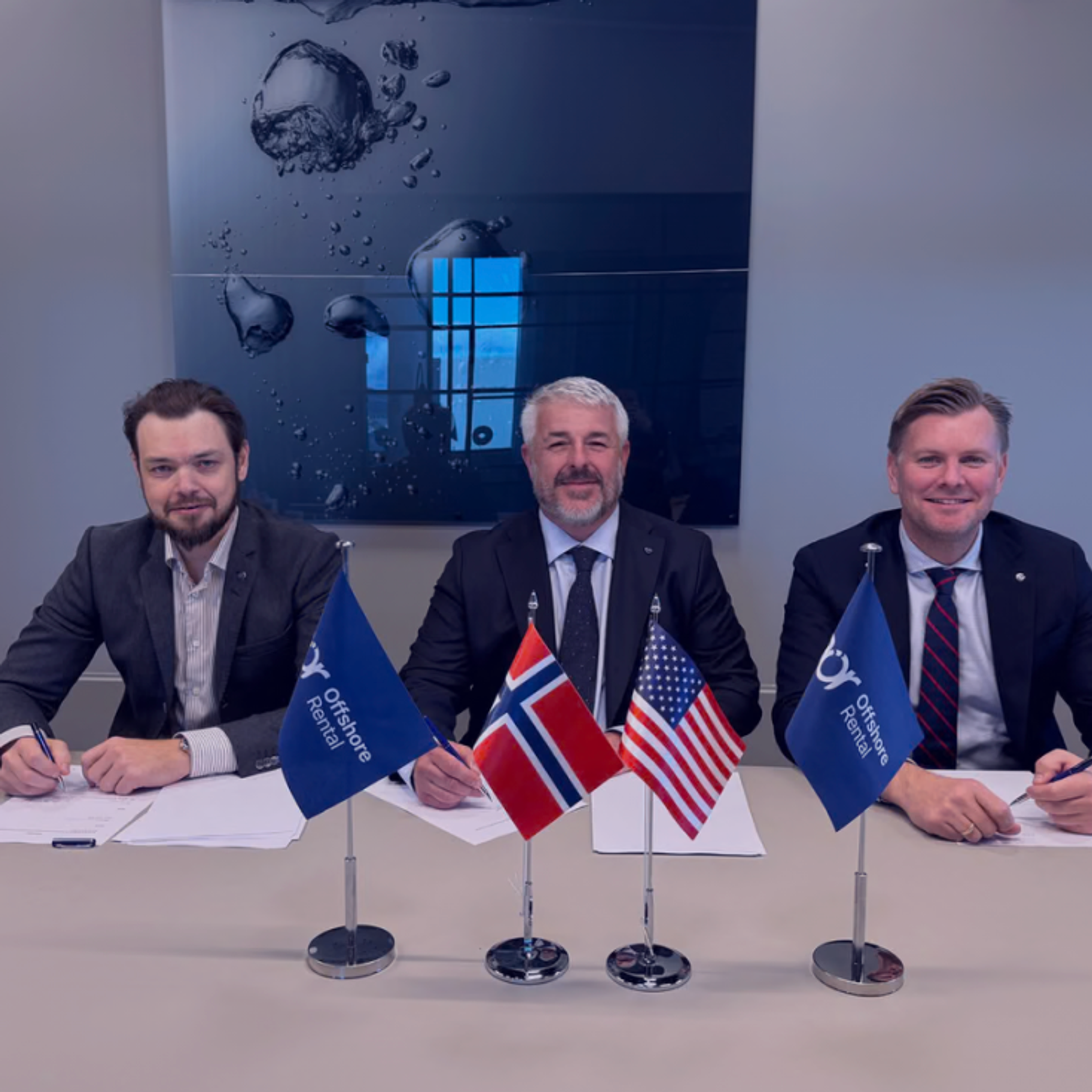 Three men in suits sit at a table with flags for Offshore Rental, Norway, and the USA, appearing to sign documents.