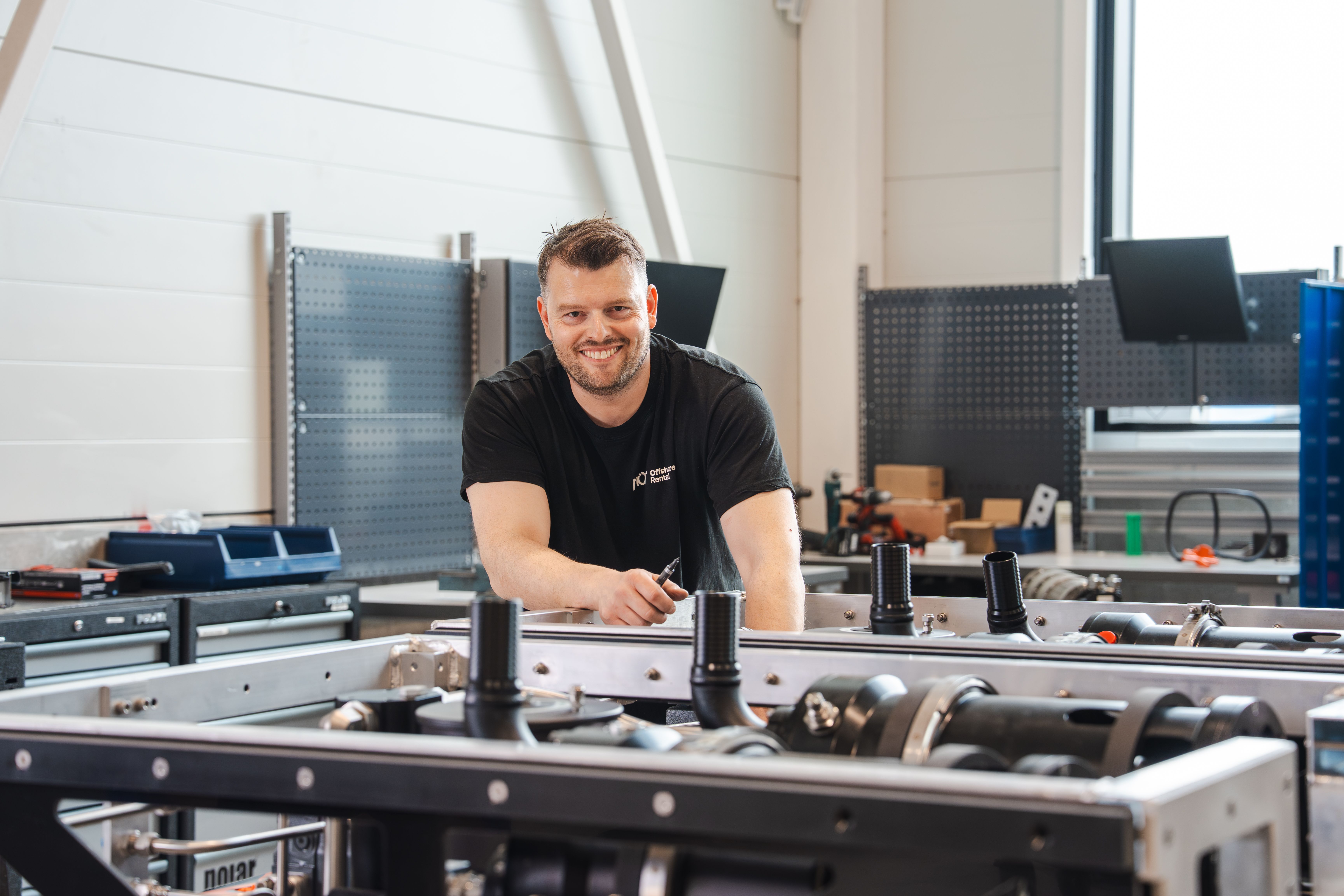 A smiling man in a black t-shirt leans over machinery in a workshop, holding a tool.