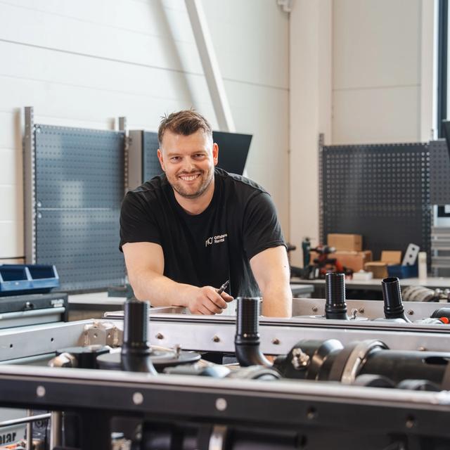 A smiling man in a black t-shirt leans over machinery in a workshop, holding a tool.