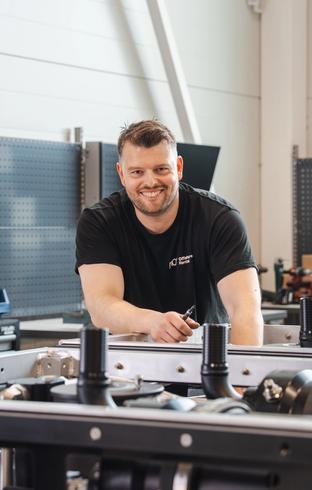 A smiling man in a black t-shirt works on machinery in a workshop.
