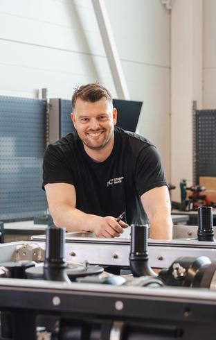 A smiling man in a black t-shirt leans on industrial machinery in a workshop, holding a tool.